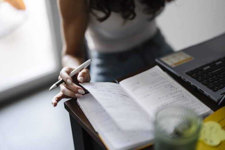 Hand writing in paper notebook at a meeting table with blurred laptop behind